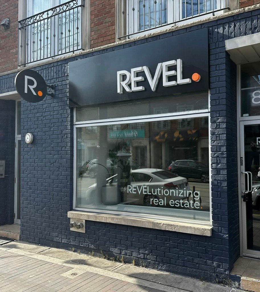 Dark navy brick storefront with illuminated 3D REVEL lettering, round blade sign and window vinyl